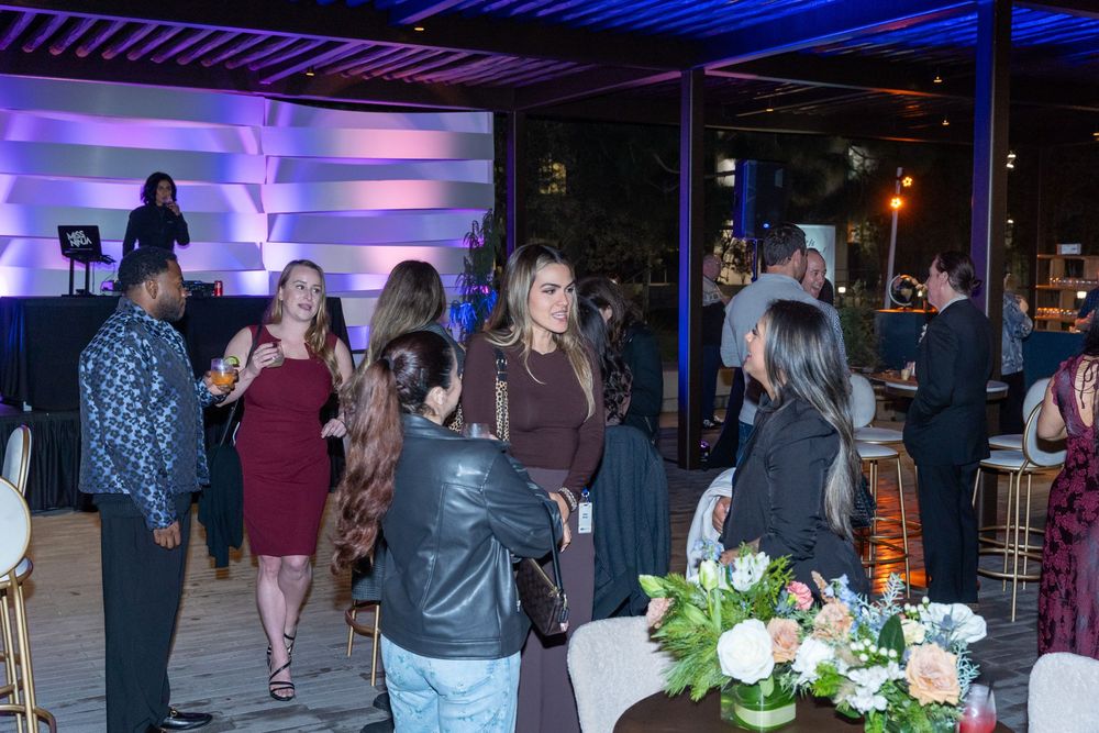 Guests mingle at an elegant evening cocktail party with purple ambient lighting and floral centerpieces.