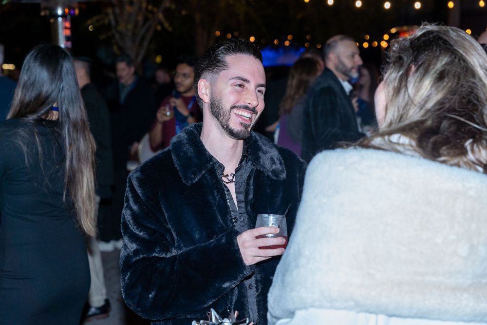 Man in black fur coat smiling and holding a drink at an outdoor evening social gathering event.