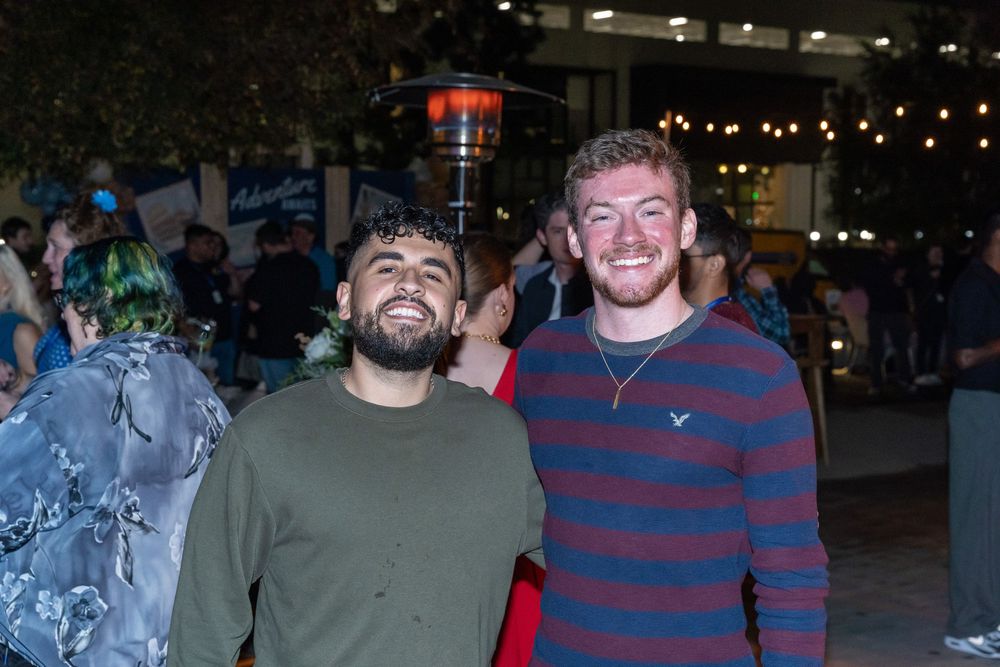 Two smiling young men posing together at an outdoor evening social event with patio heaters in background.
