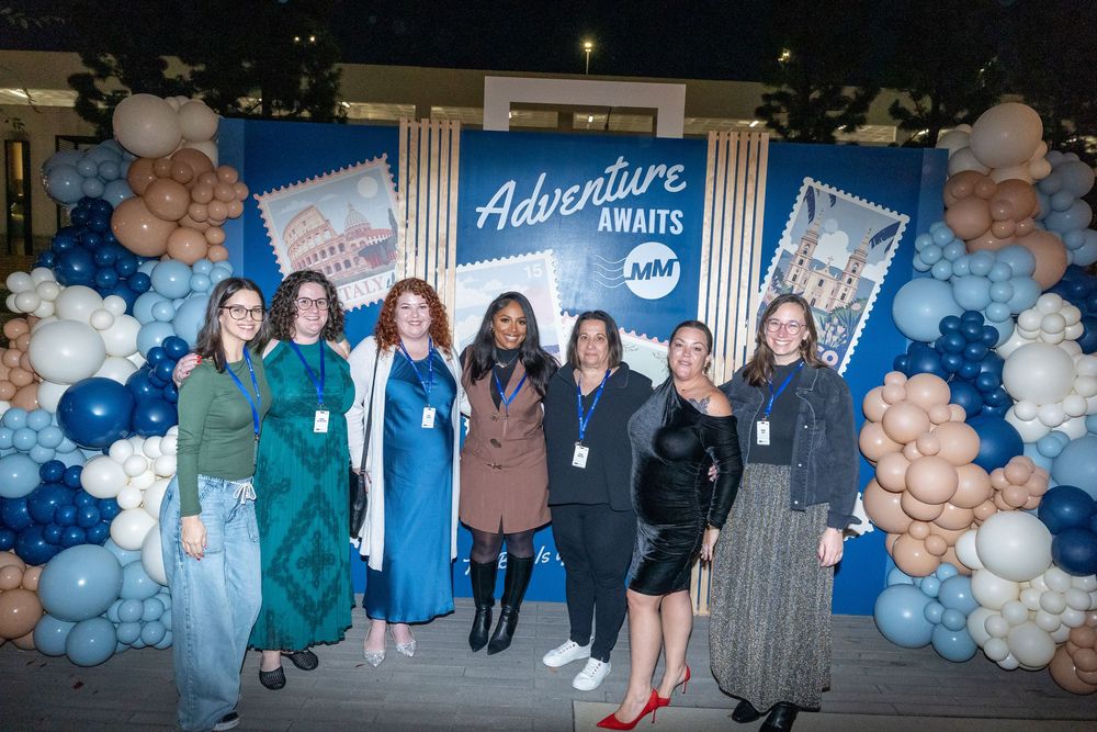 Group of seven women posing in front of an Adventure Awaits balloon backdrop at a corporate event.