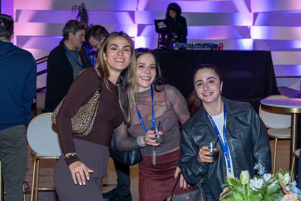 Three women smiling and posing together at an evening corporate event with purple ambient lighting.