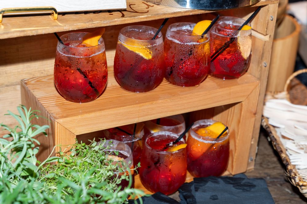 Wooden crate display holding six stemless glasses of red sangria garnished with orange slices and fresh fruit.