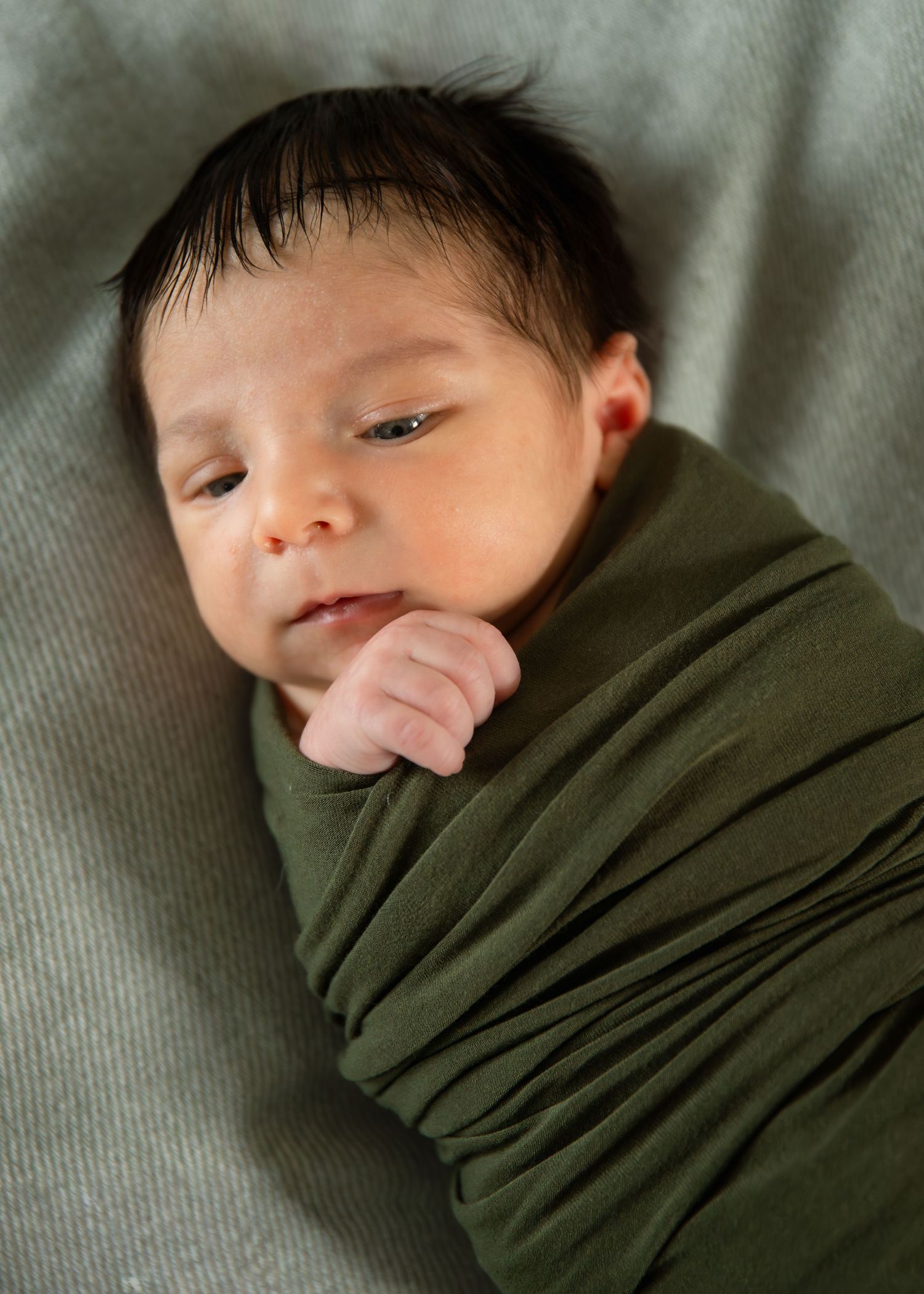 Newborn baby swaddled in olive green wrap with dark hair, resting peacefully on gray background.