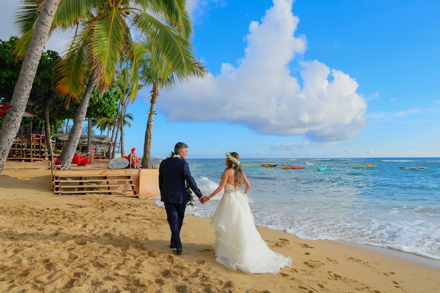 Bride and groom walk hand in hand along a tropical beach with palm trees and turquoise ocean waves.