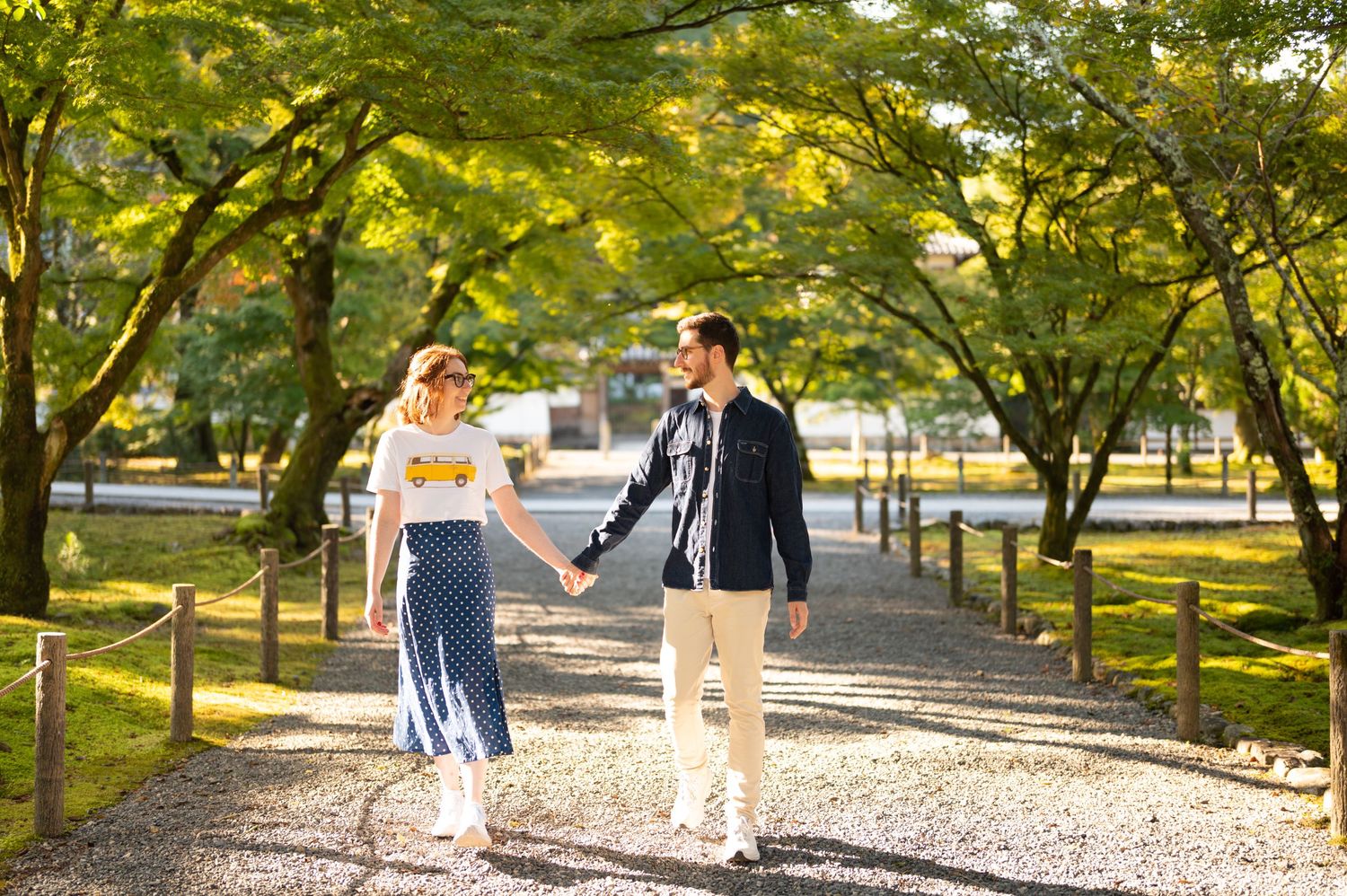 Couple holding hands walking along a tree-lined gravel path in a lush Japanese garden.