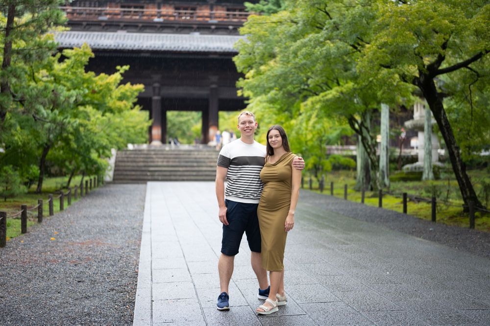 Couple posing on a stone path at a traditional Japanese temple surrounded by lush green trees.