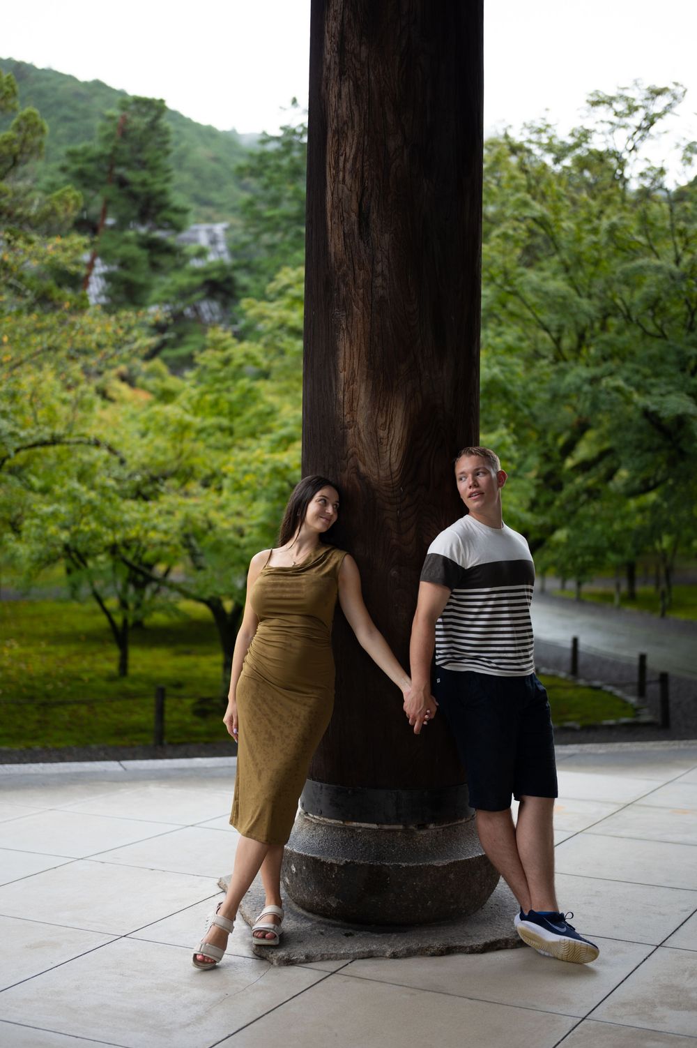 Couple holding hands leaning against a large wooden pillar in a Japanese garden setting with lush green trees.