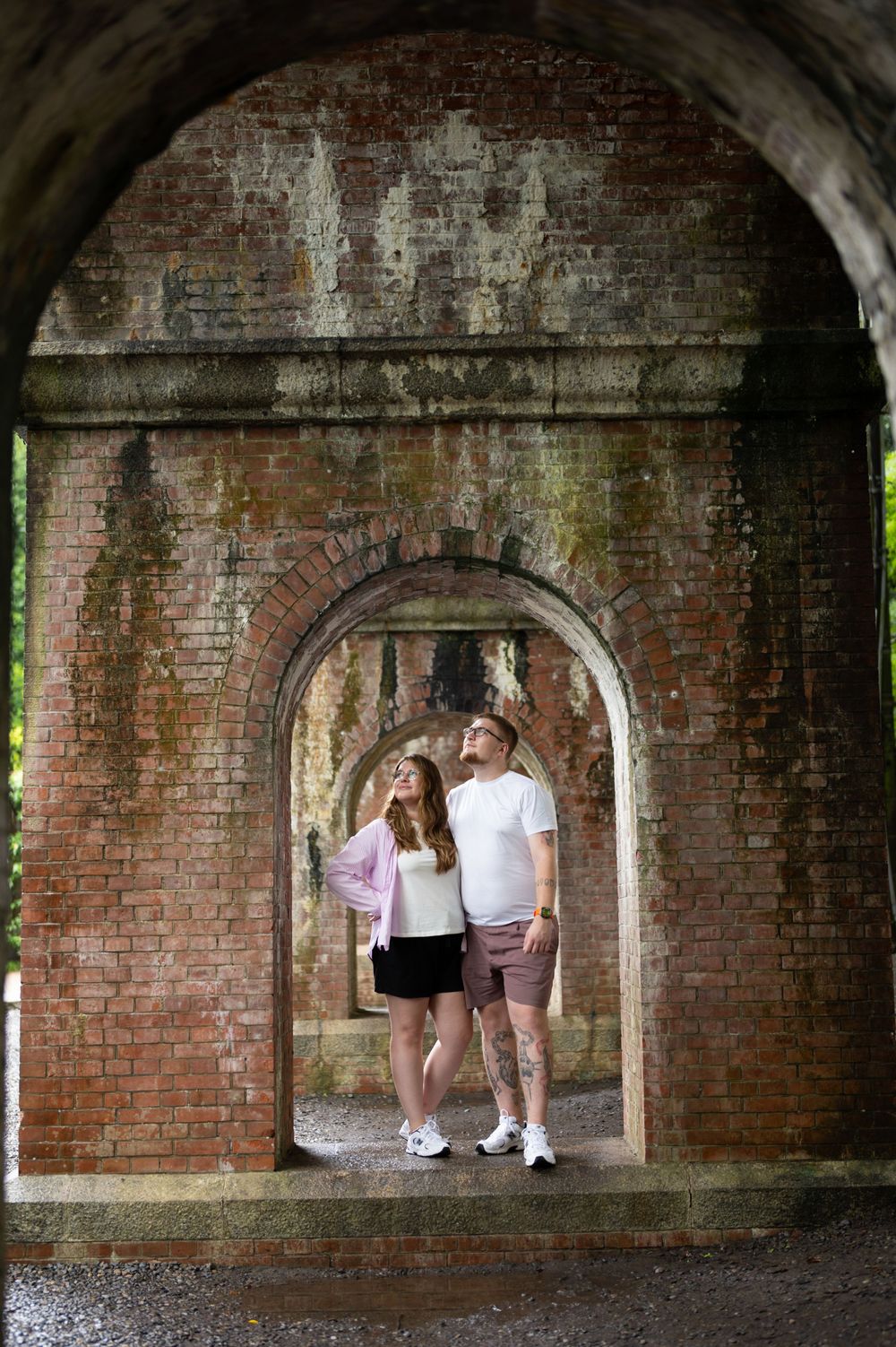 Couple standing under aged brick archway, exploring historic stone tunnel architecture together.