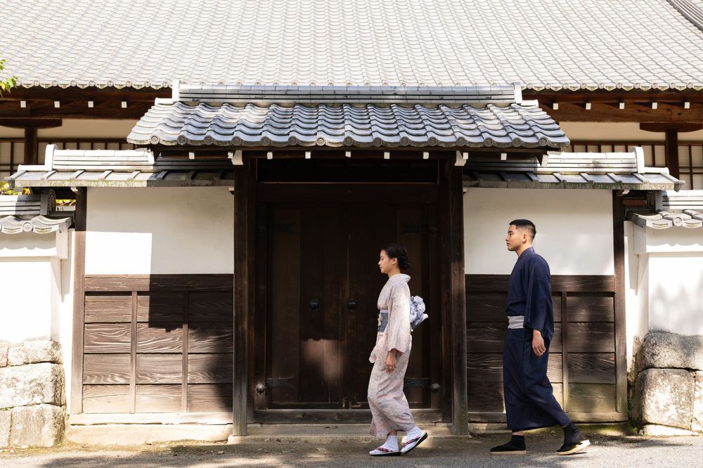 Couple wearing traditional Japanese kimono walking past a historic Japanese building with white walls and tiled roof.