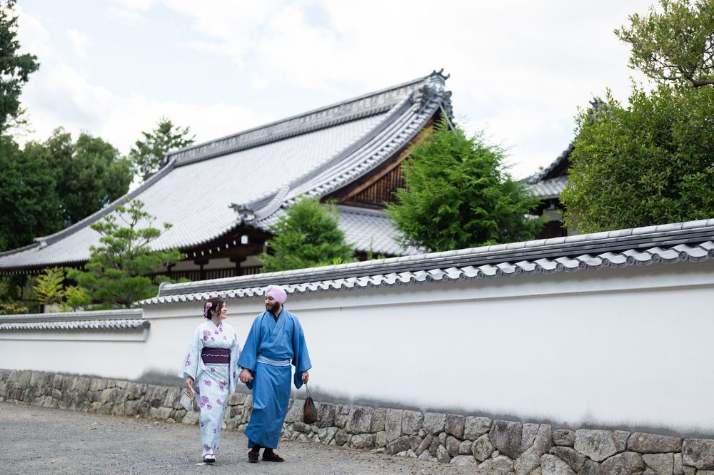 Two people in traditional Japanese kimono walk past a white stone wall and temple building in Kyoto, Japan.