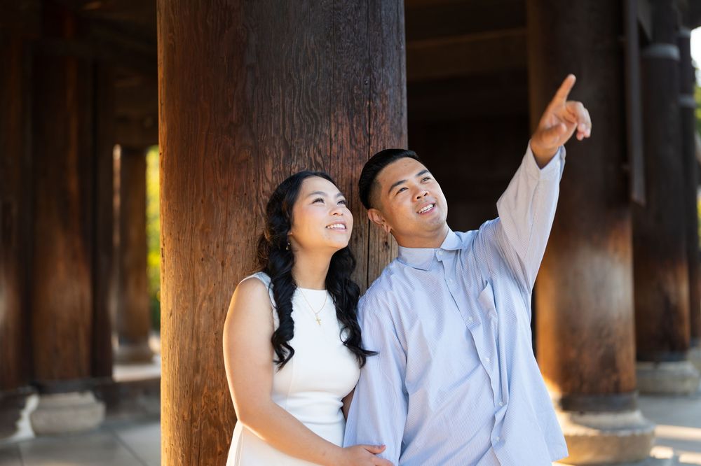 Happy Asian couple smiling and pointing at something, standing between wooden columns outdoors.
