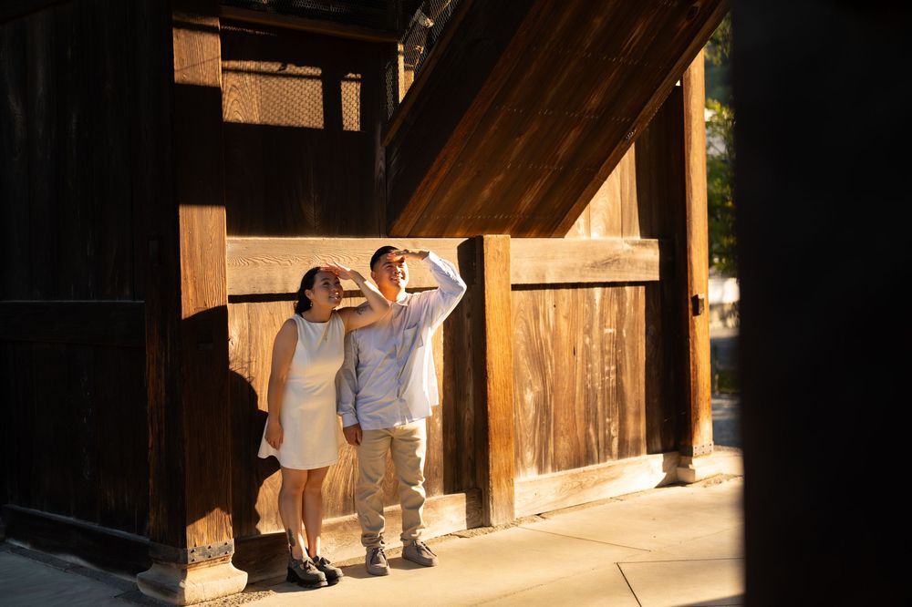 Couple shielding eyes from golden sunlight beside rustic wooden building exterior.