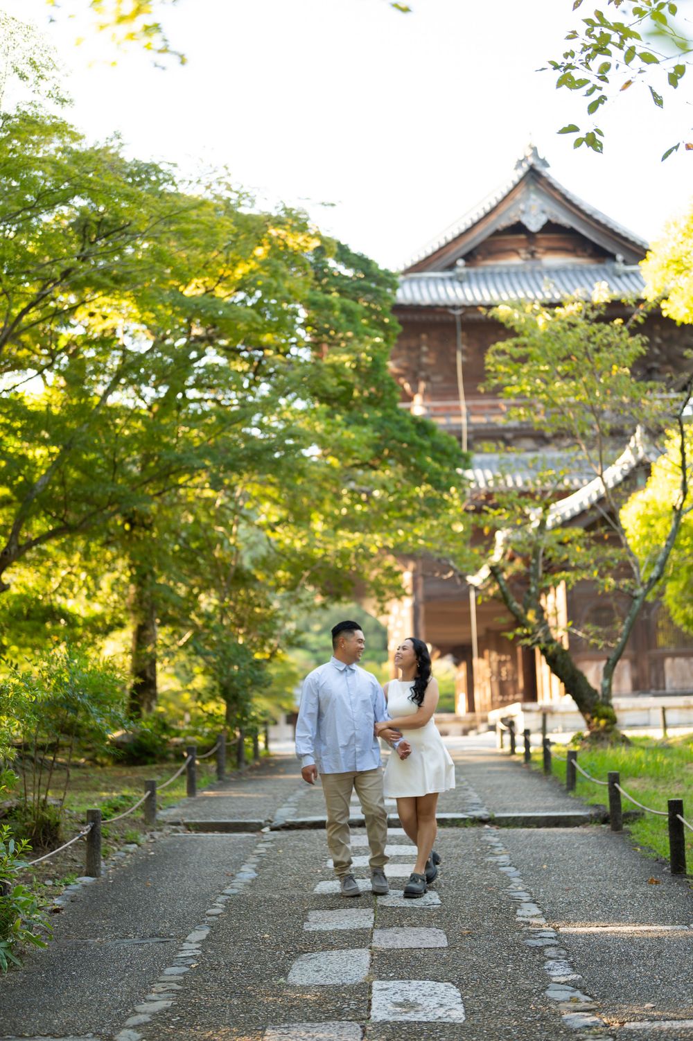 Couple holding hands walking along a stone path in a lush Japanese garden with a traditional wooden temple.