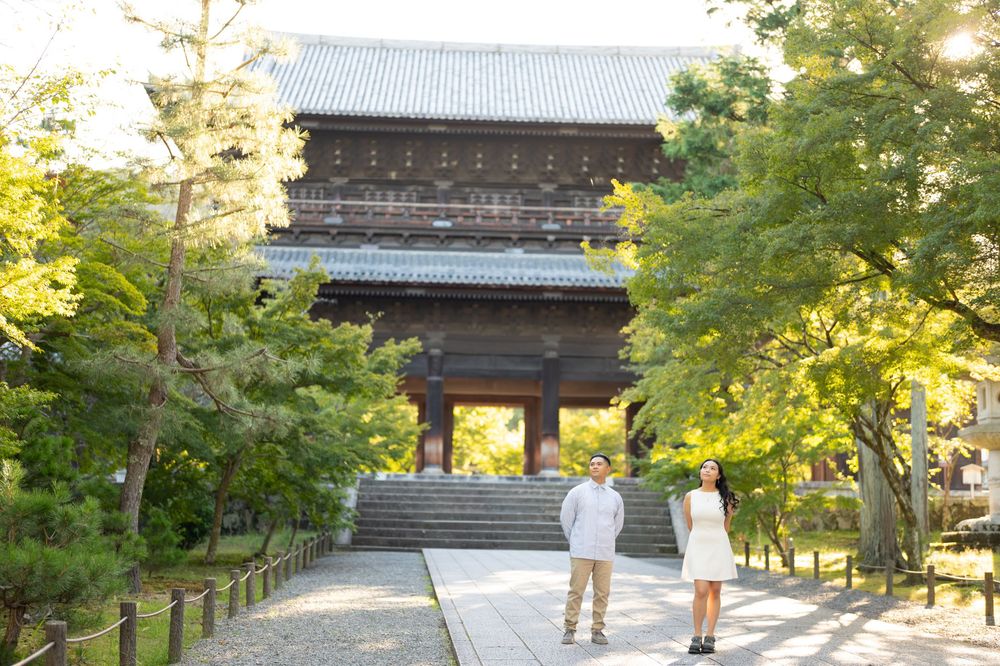 Couple standing on a stone path before a historic Japanese temple gate surrounded by lush green trees.