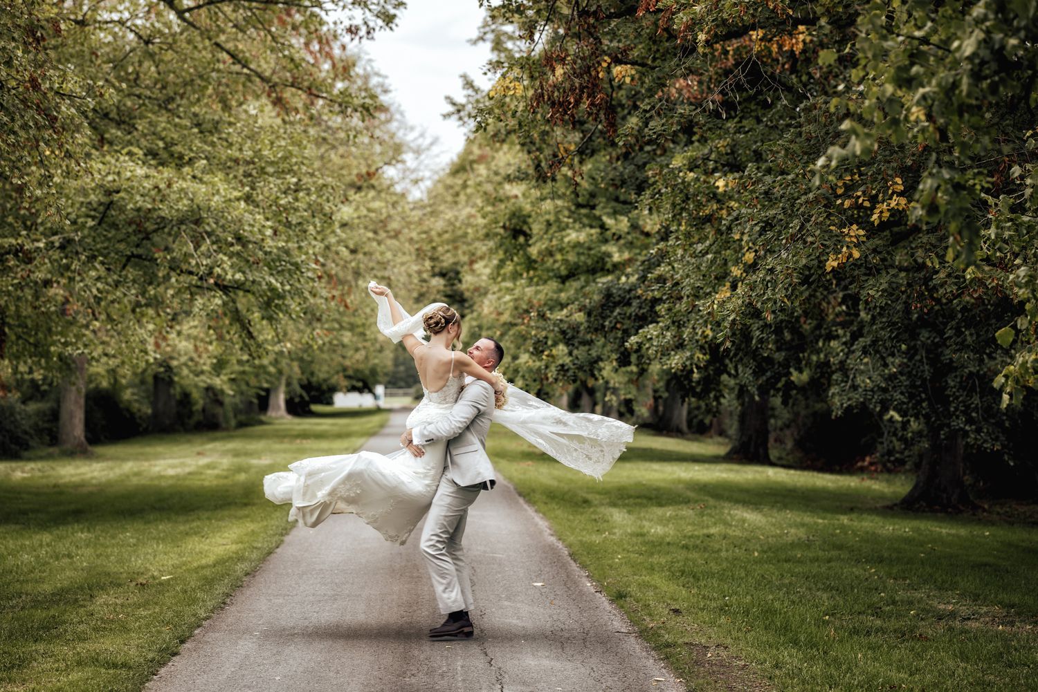 Groom lifts bride in white gown on tree-lined path in lush green park during outdoor wedding celebration.