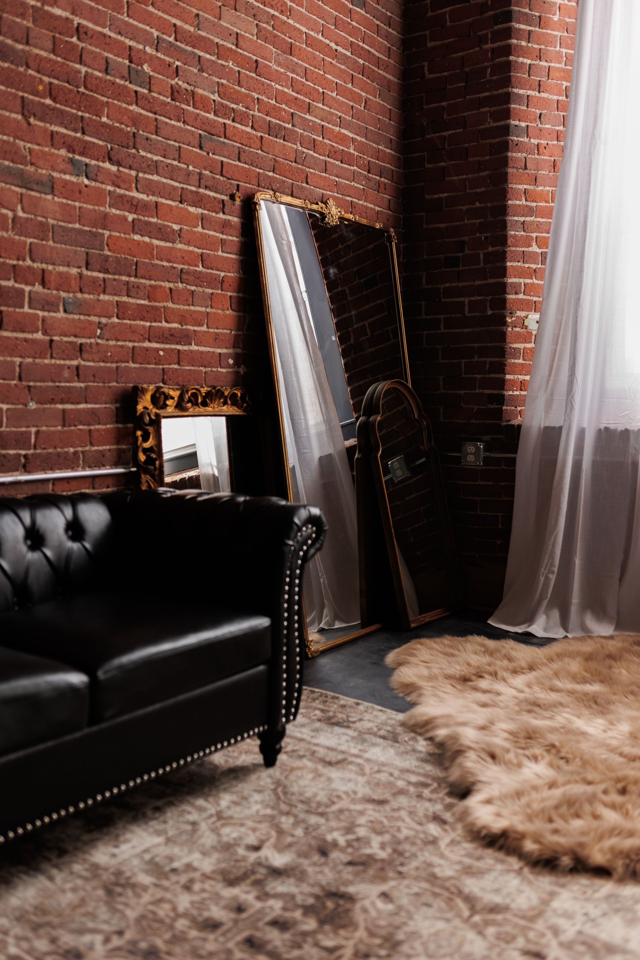 Black leather tufted sofa against exposed brick wall with mirror and furry rug in modern industrial room.