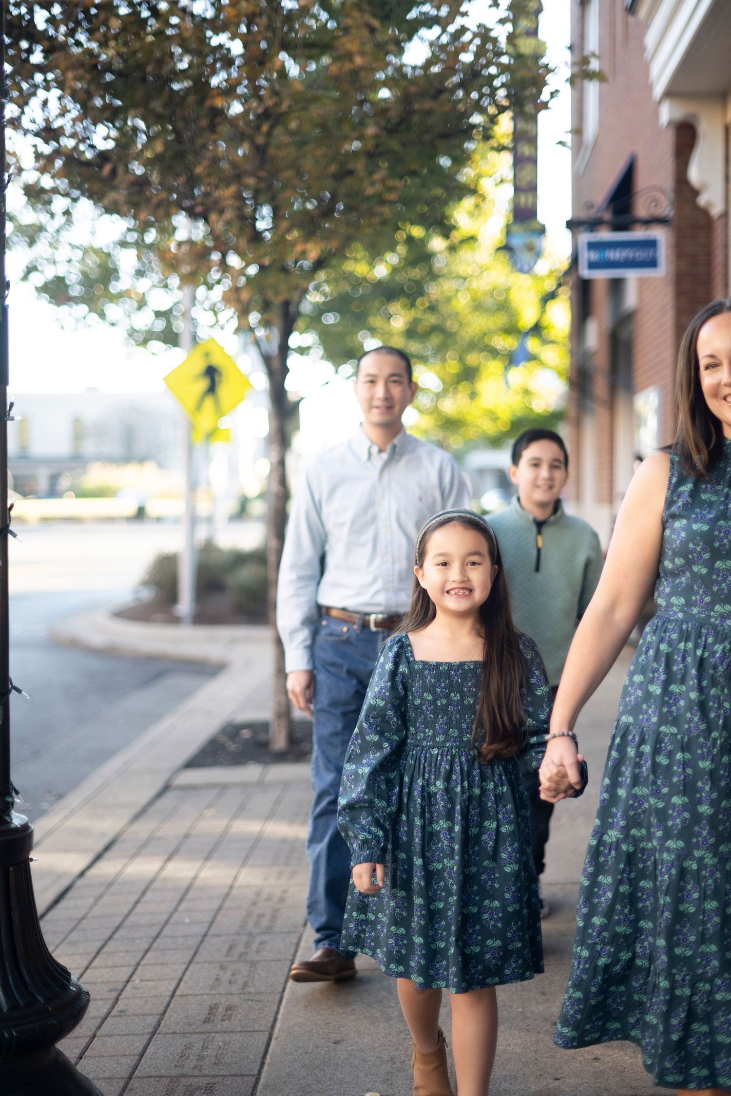 A smiling Asian family walks along a sunny urban sidewalk, with a young girl in a floral dress holding hands with her mother.