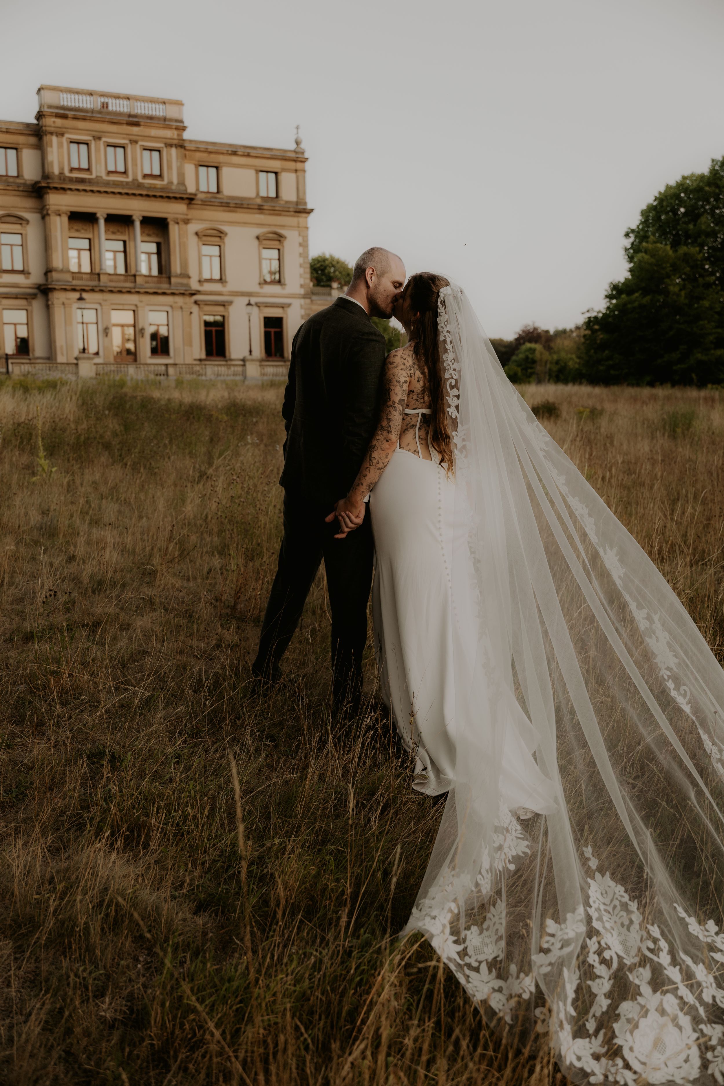 A bride and groom share a kiss in a field before a grand estate, her lace veil flowing dramatically in the breeze.