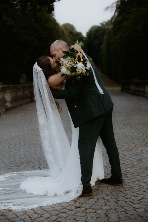 Bride and groom share a romantic dip kiss on a cobblestone path, bride holding a sunflower bouquet in a white gown.