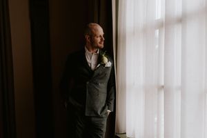 Groom in a dark suit with white boutonniere stands by a sunlit curtained window, gazing thoughtfully to the side.