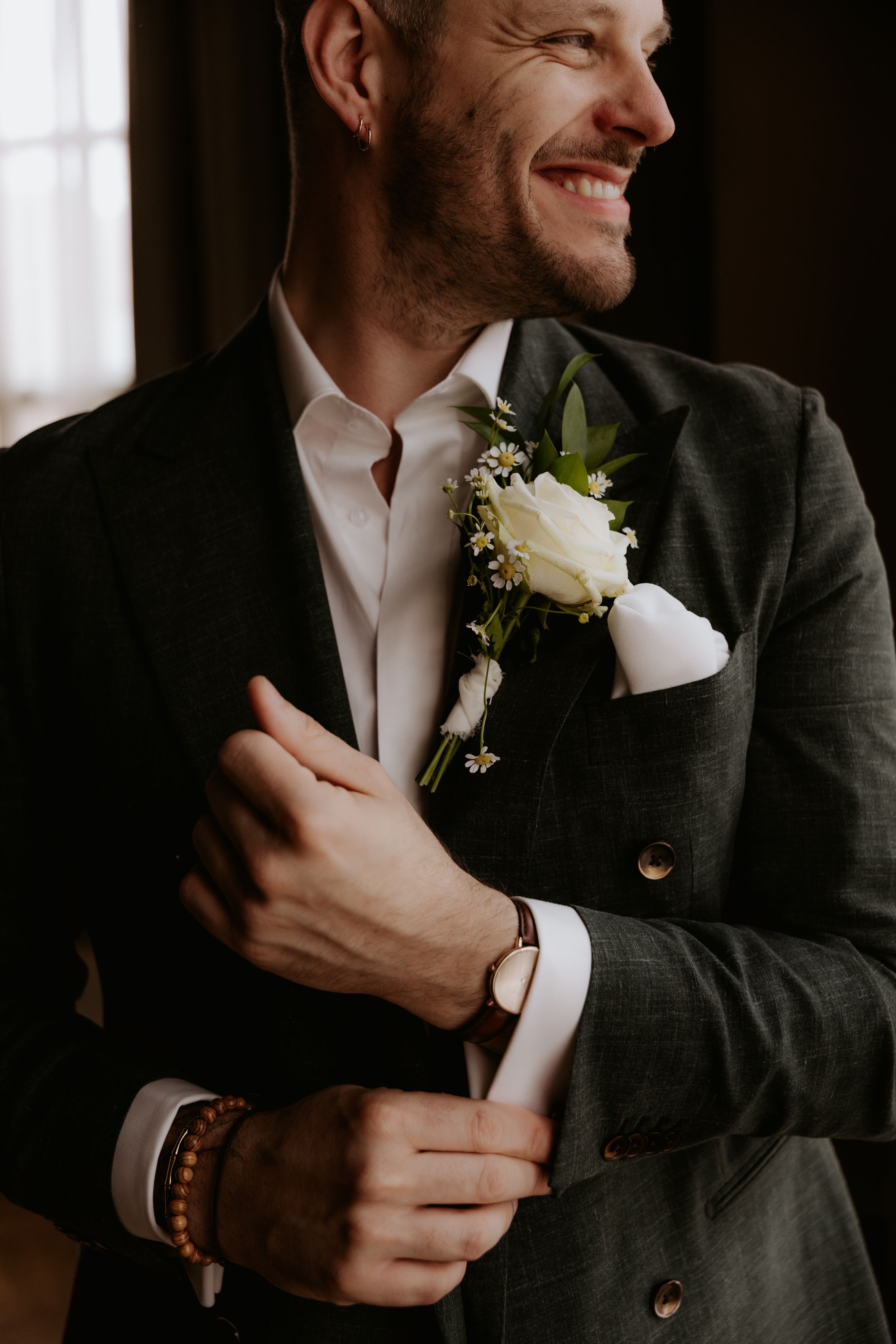 Smiling groom in dark suit adjusting cufflinks with white rose boutonniere and pocket square on wedding day.