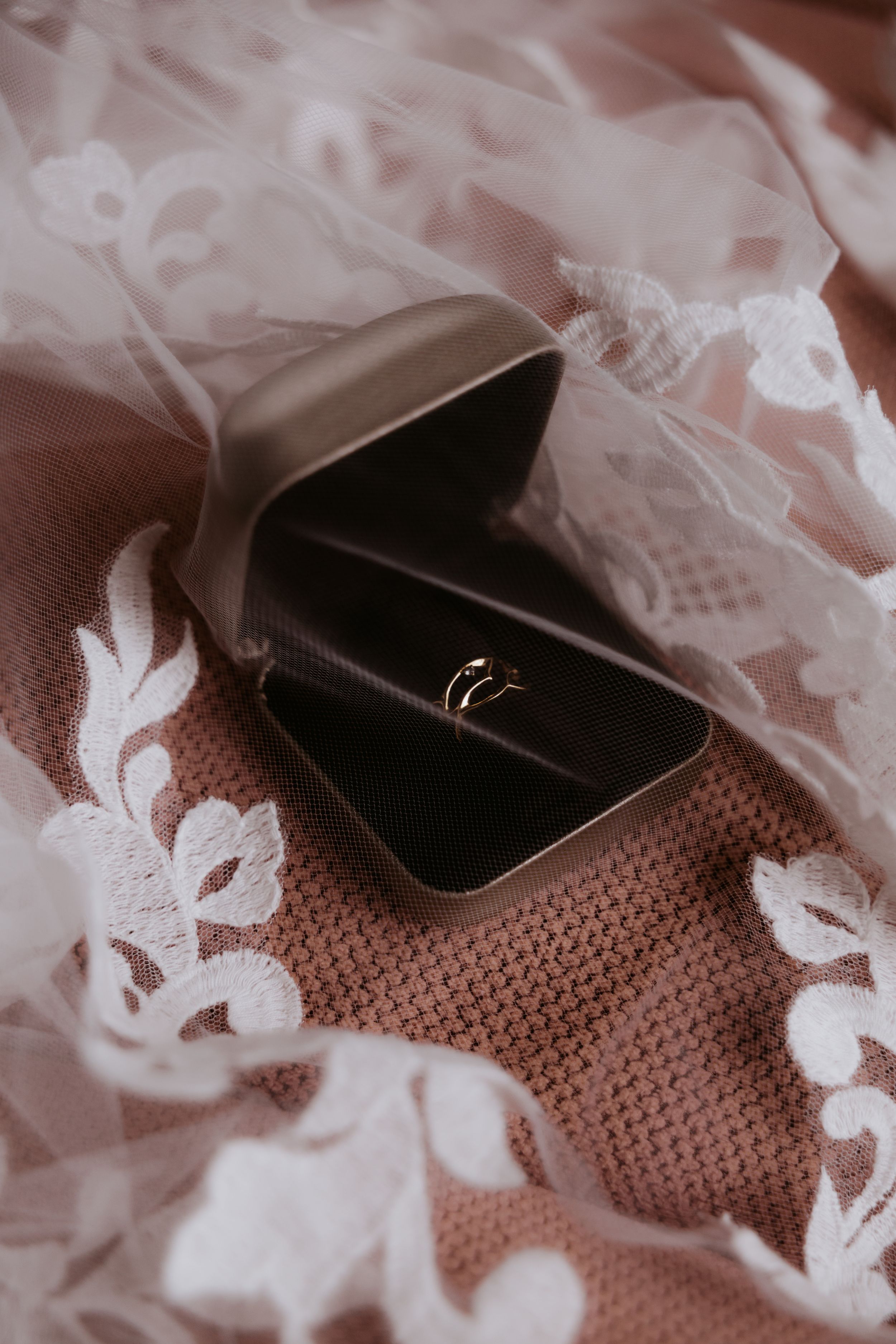 Gold wedding rings in a dark jewelry box on a pink knitted fabric with white lace wedding veil.