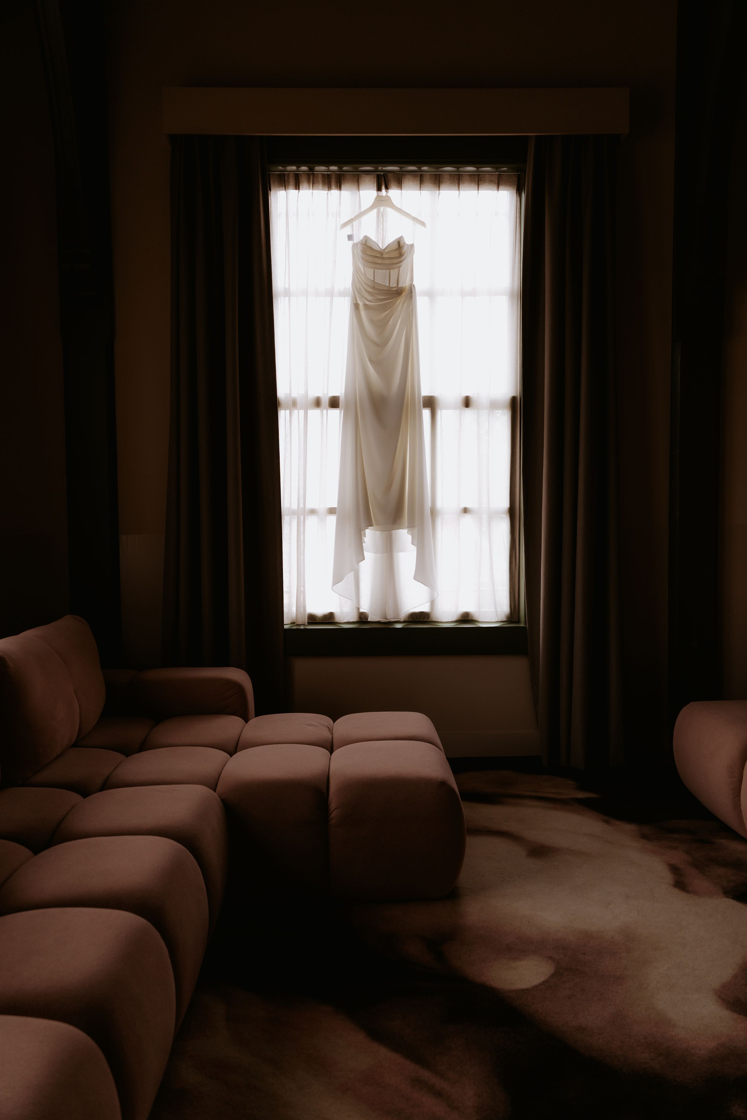 A wedding dress hanging in a sunlit window, backlit by natural light, with dark curtains and a modern sofa in a dim room.