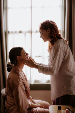 A makeup artist with curly red hair applies makeup to a smiling bride in a satin robe near a bright window.