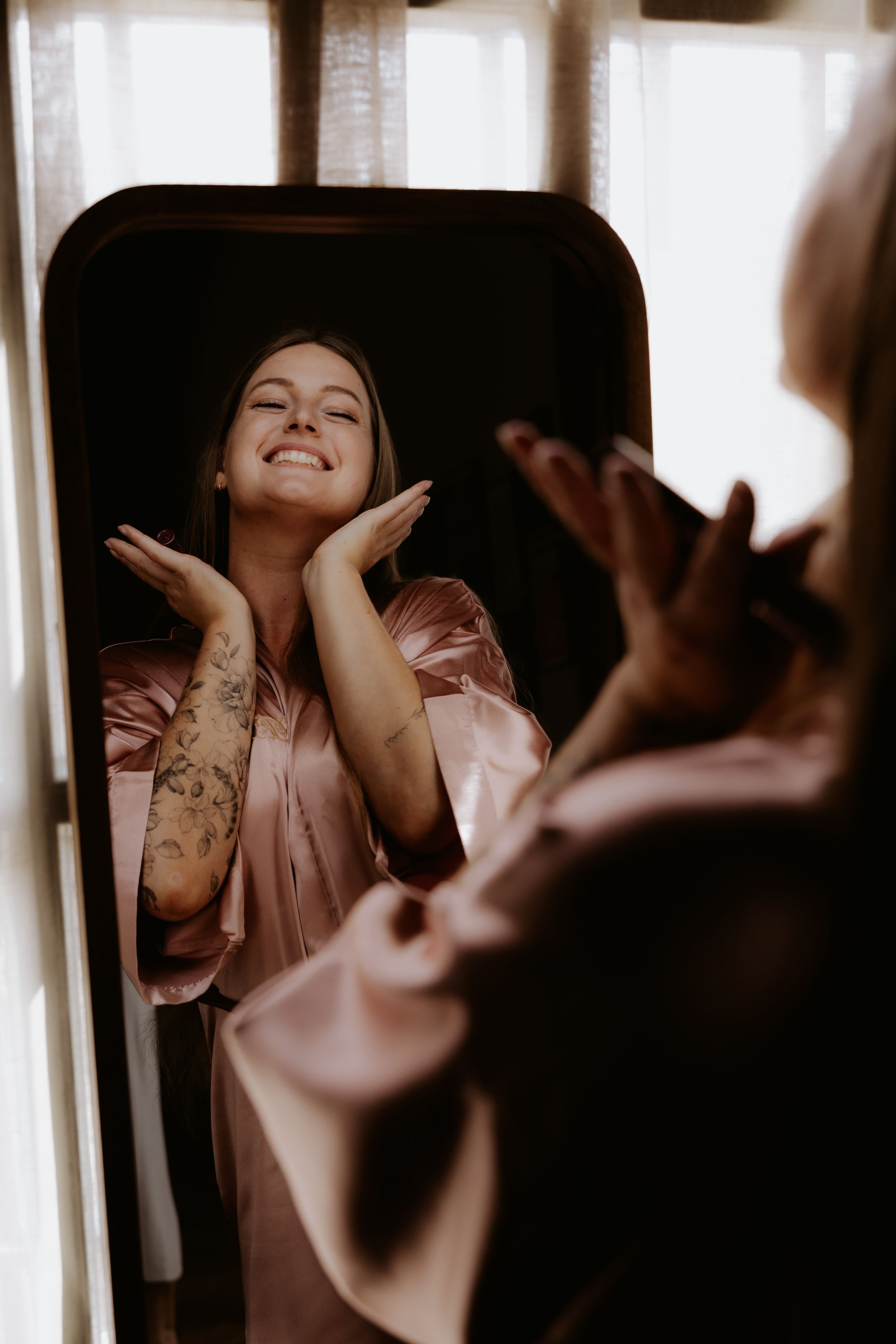 A smiling woman in a pink satin robe with tattoos poses in front of a mirror, hands framing her face.