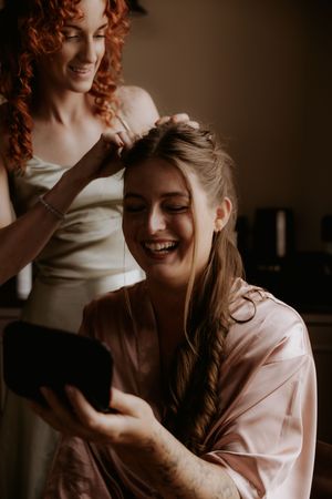 Bride smiling at mirror while bridesmaid with red curly hair styles her hair, both wearing satin robes.