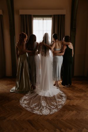 Bride in lace veil with bridesmaids in sage and black gowns standing together facing window on wedding day.