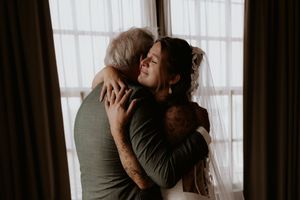 A bride in a veil shares a tender embrace with an older man near a bright window on her wedding day.