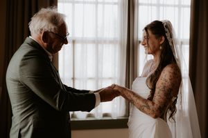 Bride in white gown and veil holds hands with elderly man in suit near window on wedding day.
