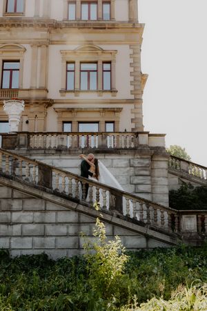 Bride and groom share a romantic kiss on grand stone staircase outside elegant historic mansion on wedding day.