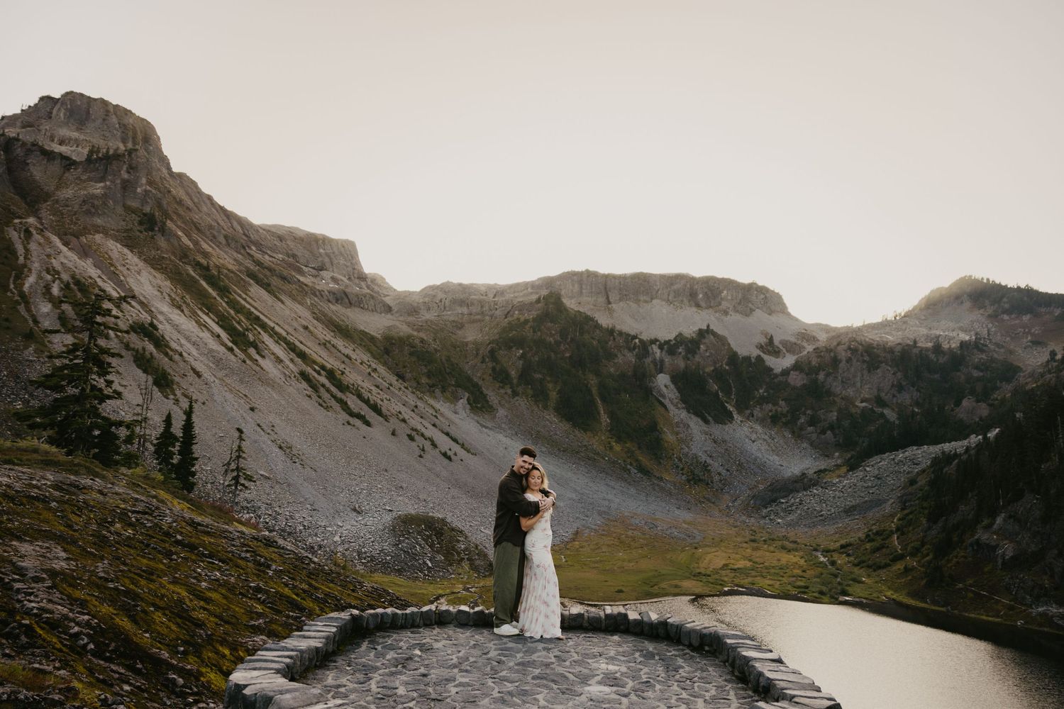 Couple embraces on rocky mountain summit with alpine lake and dramatic peaks in background during outdoor wedding elopement North Cascades