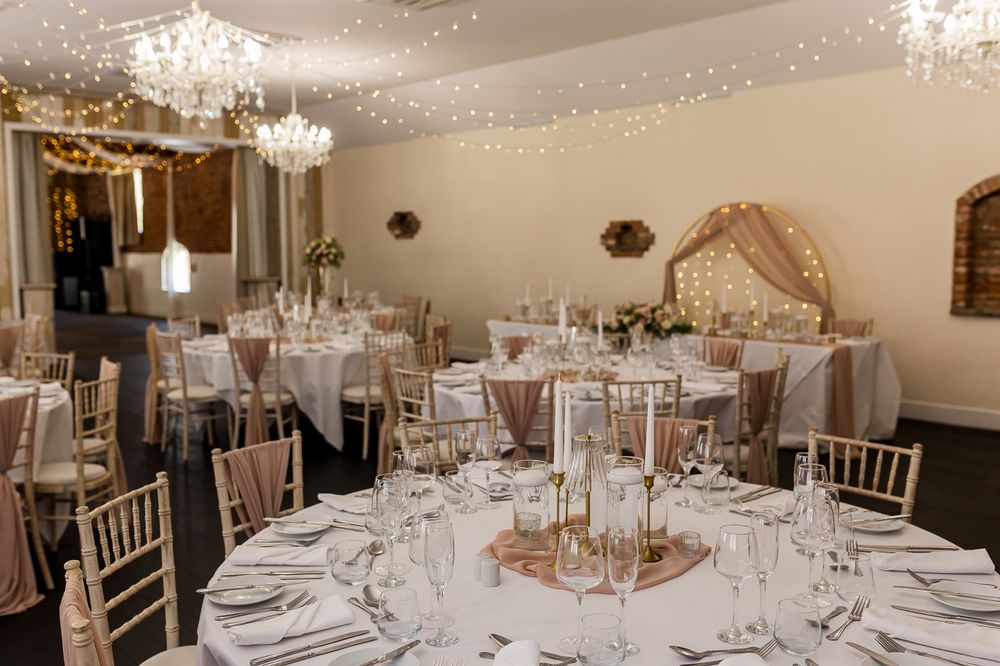 Elegant wedding reception room with fairy lights, chandelier, gold chiavari chairs, and white-clothed tables with candle centerpieces.