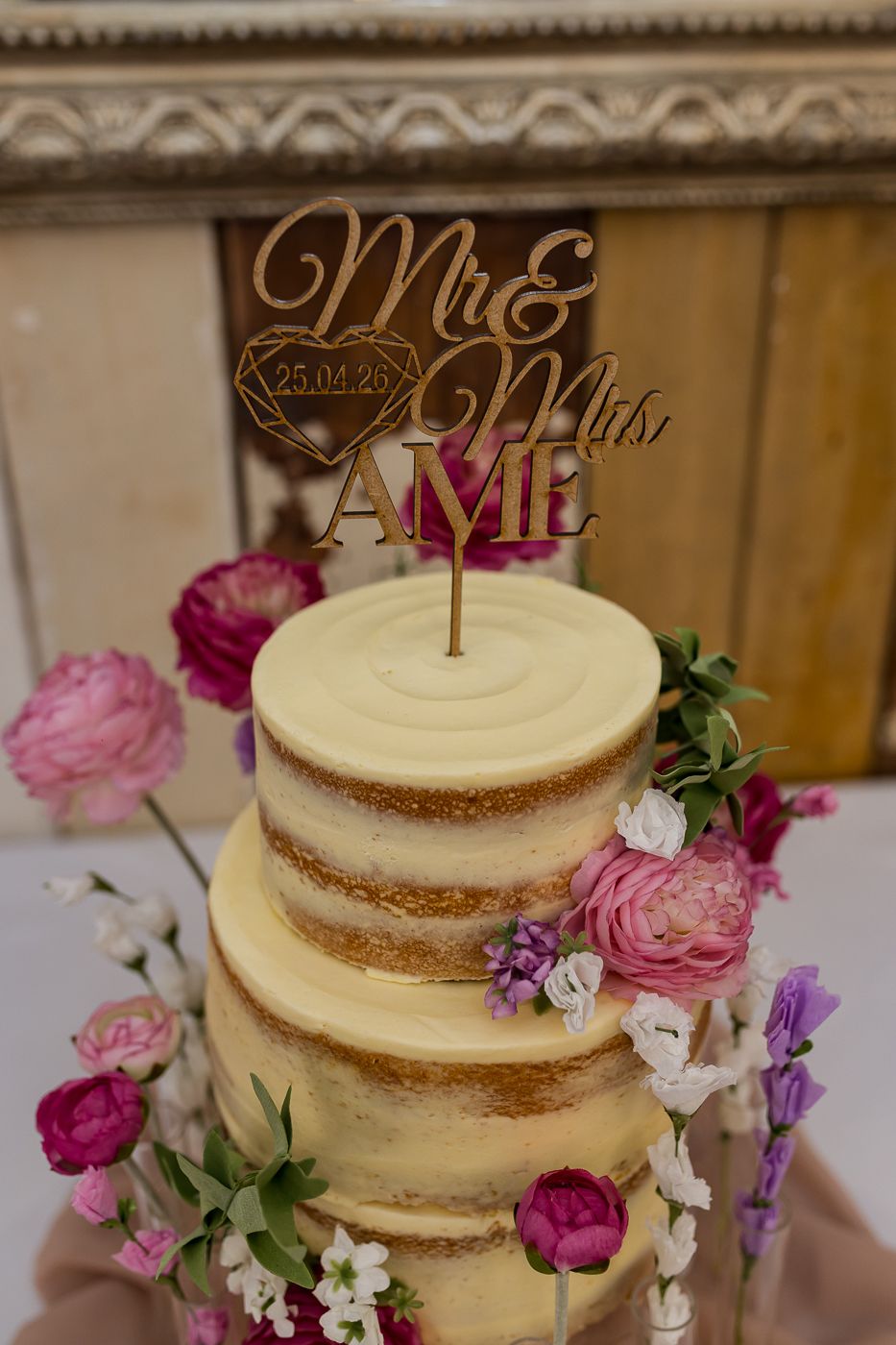 Two-tier naked wedding cake with pink floral decorations and a gold Mr & Mrs cake topper on a wooden table.