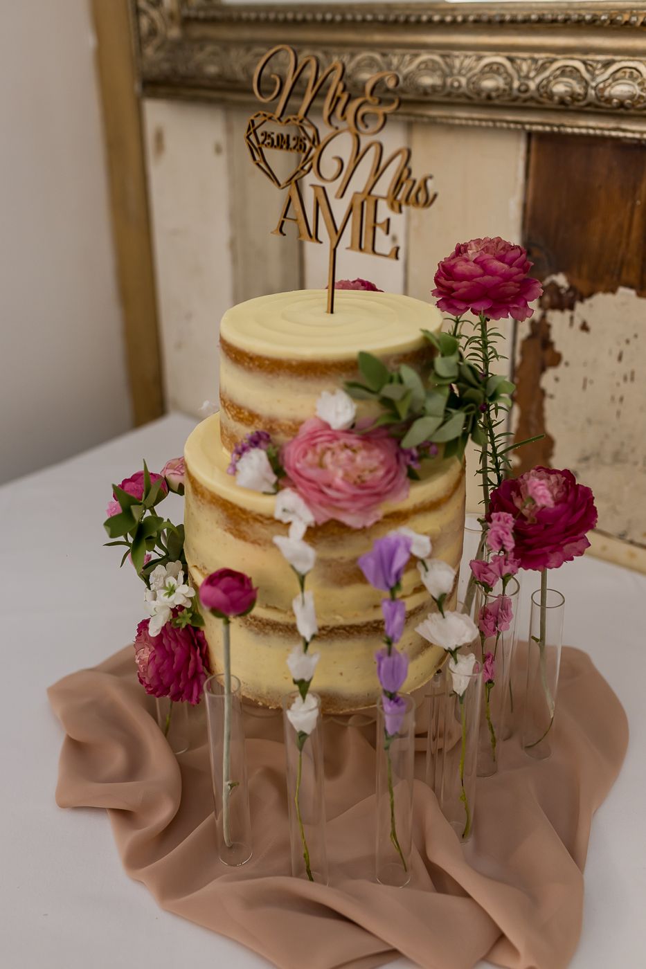 Two-tier naked wedding cake with pink flowers and a Mr & Mrs cake topper, displayed on a draped table.