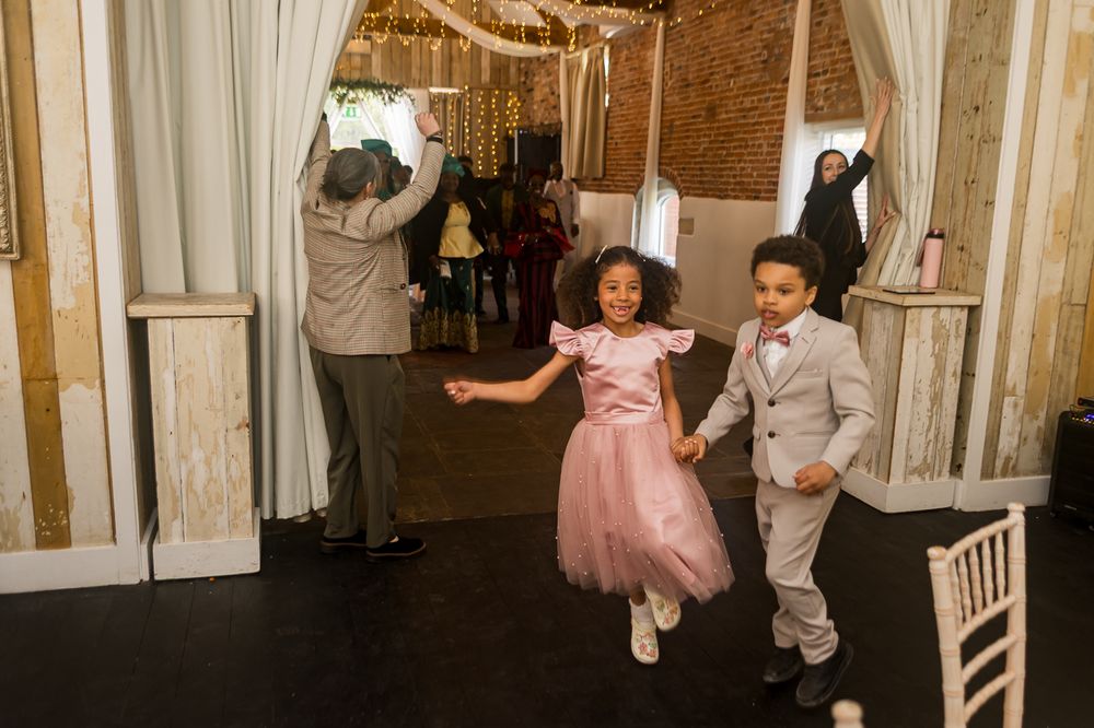 Two children in formal attire dance joyfully at a wedding reception in a rustic venue with string lights.