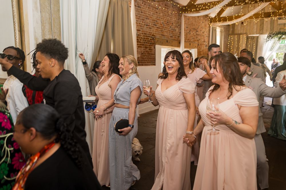 Bridesmaids in blush pink dresses laugh and dance together at a wedding reception in a rustic brick venue.