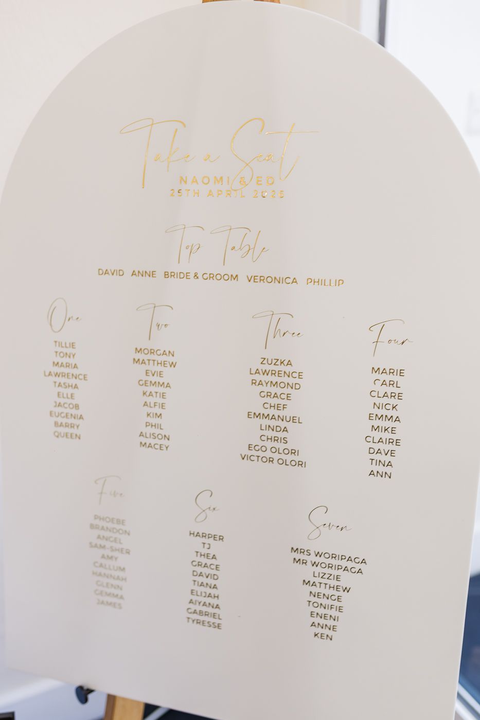 Elegant white arch-shaped wedding seating chart with gold typography displaying table assignments for guests.