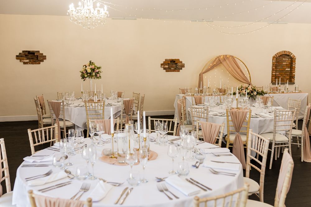 Elegant wedding reception hall with round tables, white linens, chiavari chairs, floral centerpieces, and a chandelier.