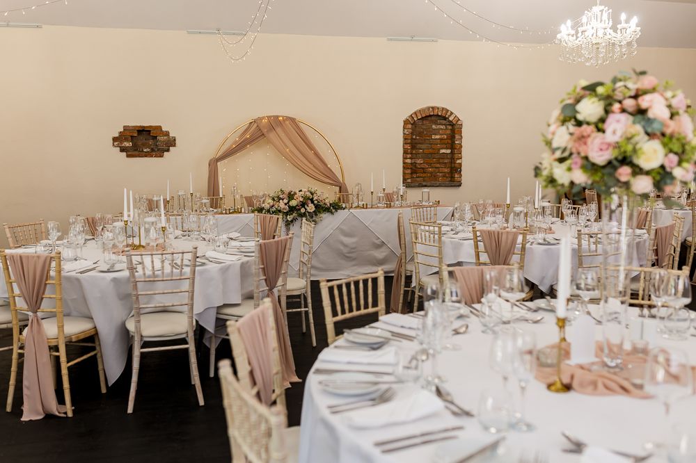 Elegant wedding reception hall with gold chiavari chairs, blush pink draping, floral centerpieces, and white round tables.