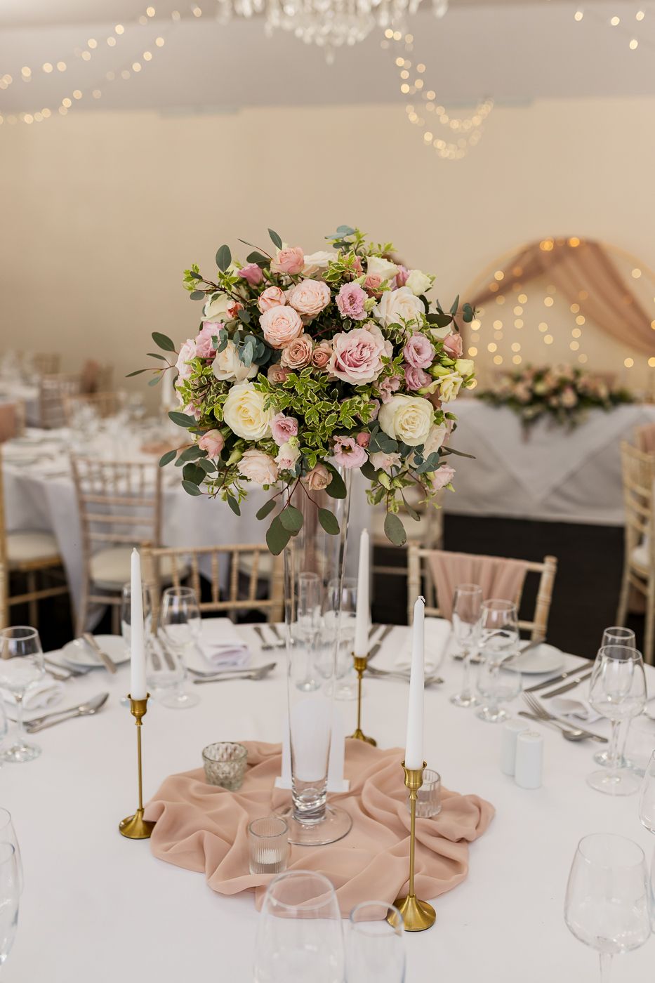 Elegant wedding reception centerpiece with pink roses in tall glass vase, gold candle holders, and blush table runner.