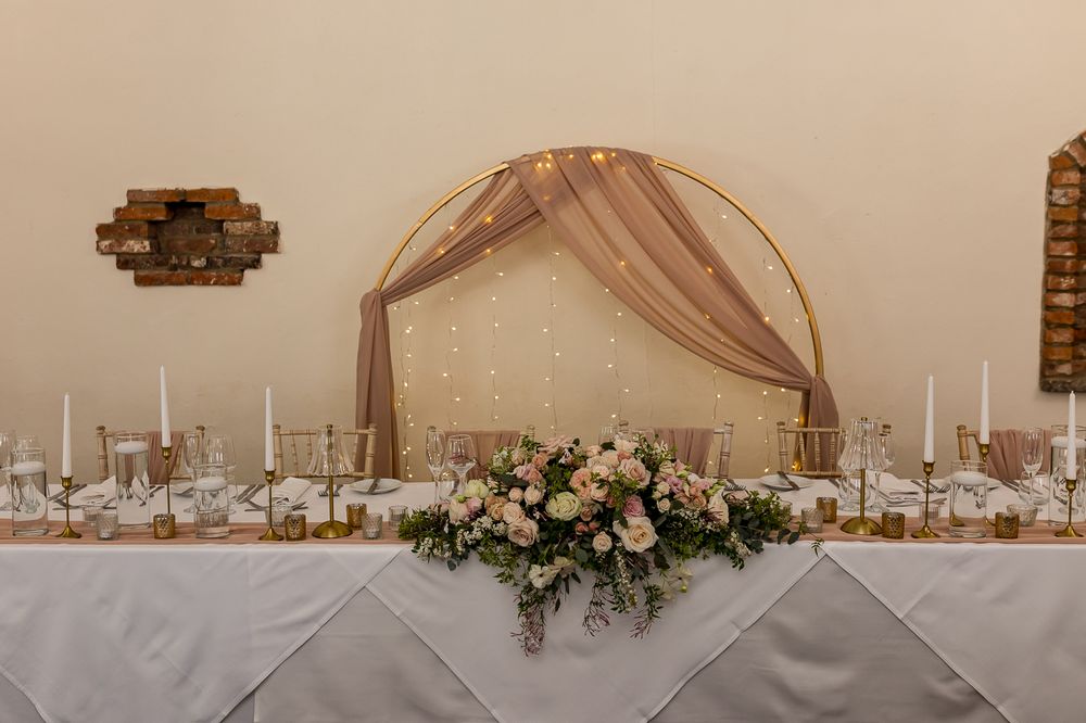 Elegant wedding head table with floral centerpiece, candles, and a golden hoop arch draped in blush fabric and fairy lights.