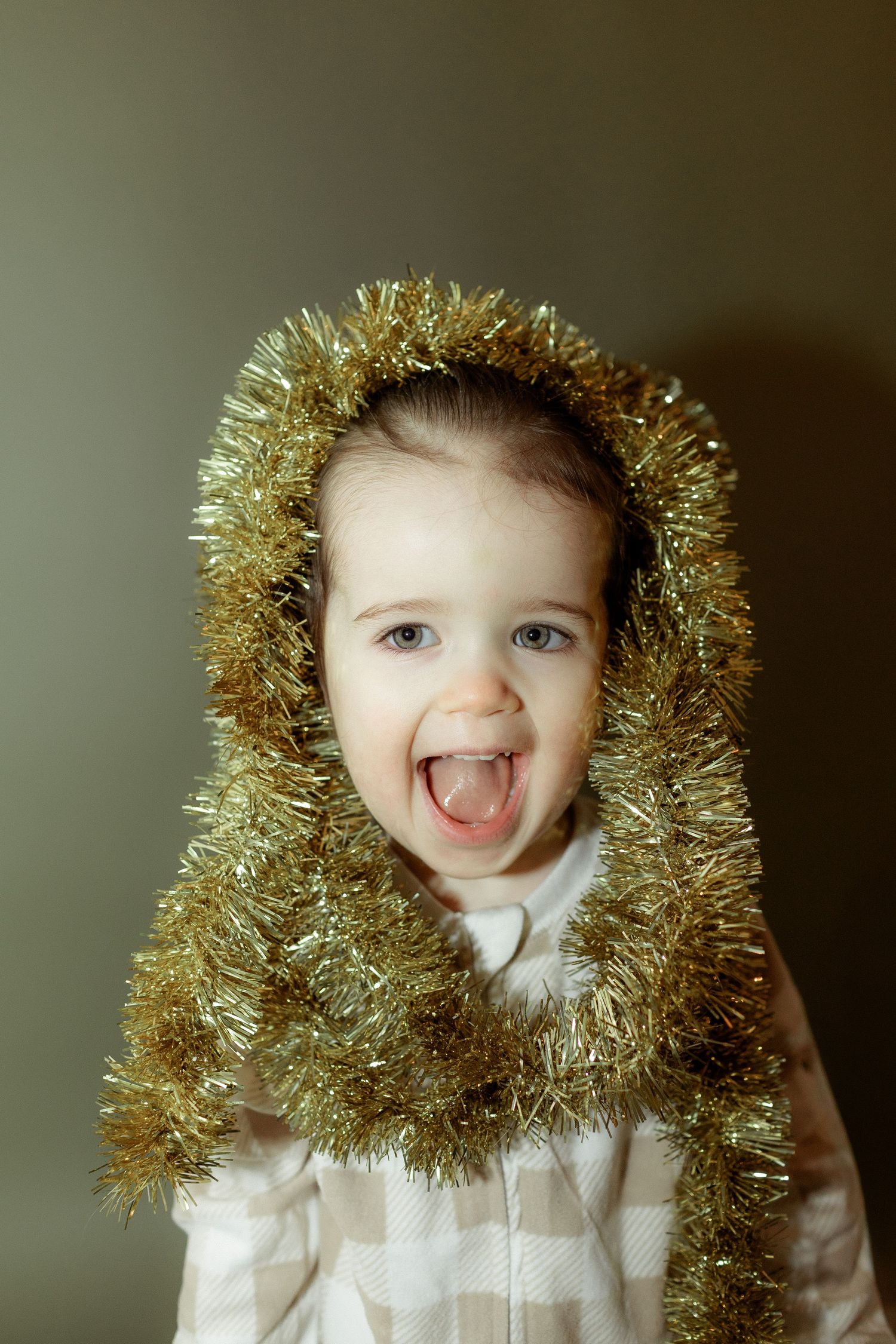 Joyful toddler wearing gold Christmas tinsel as a hood, laughing with mouth open in festive holiday portrait.