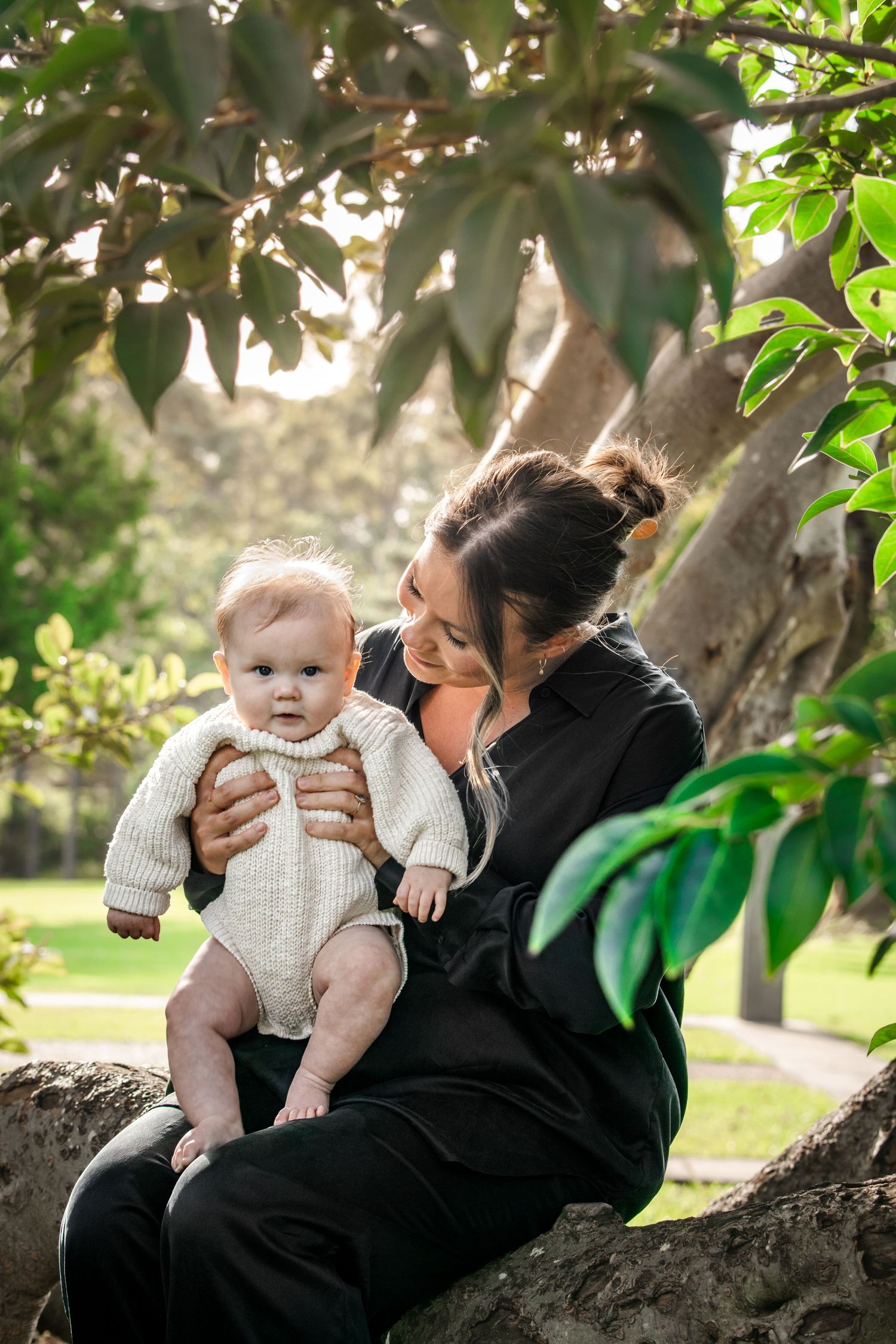 A woman in a black shirt sits on a tree branch in a sunny park, smiling and holding a baby in a cream knit sweater in Castle Hill.