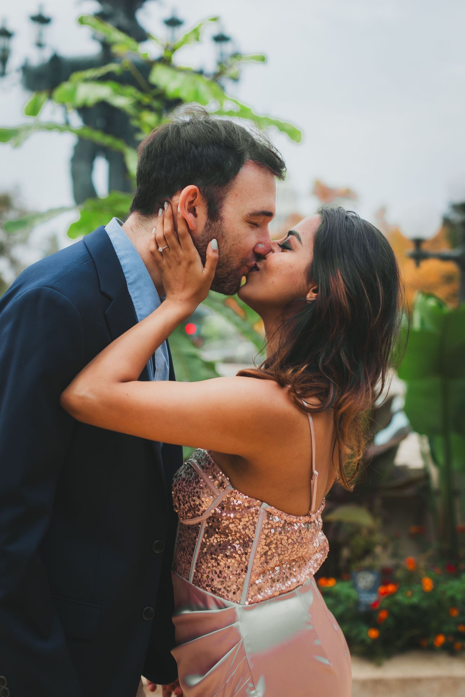 Couple sharing a romantic kiss outdoors, the woman in a sparkly rose gold dress and the man in a navy blazer, surrounded by lush greenery.