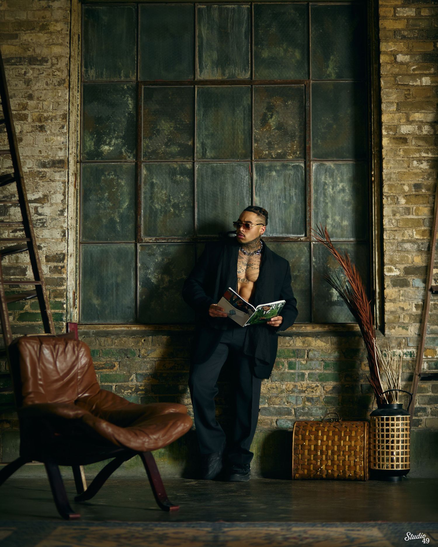 Man in dark suit holds open book against industrial brick wall with leather chair and wicker basket nearby.