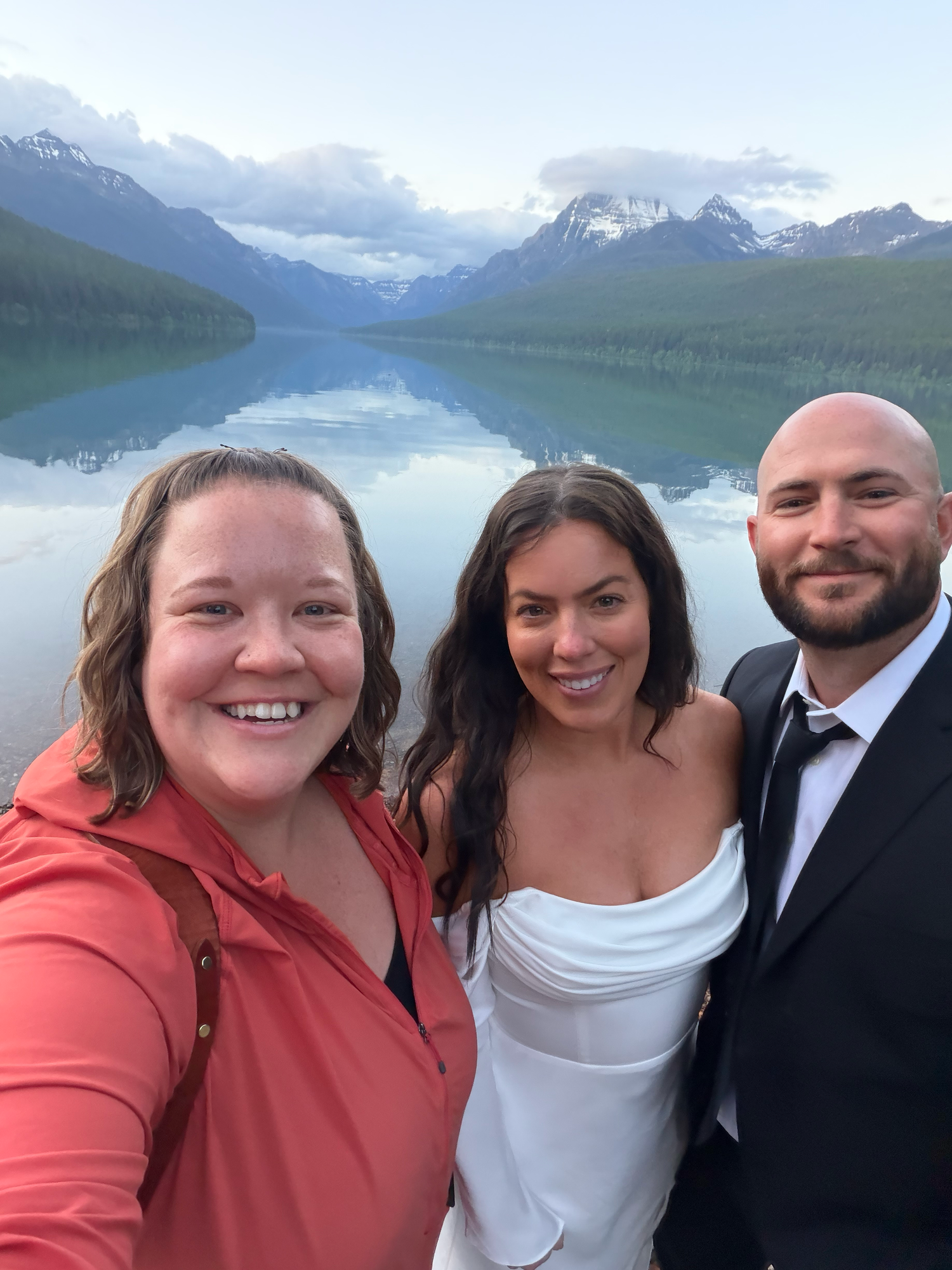 Woman in white blouse and jeans smiling on a mountain overlook with a scenic lake and forest backdrop.